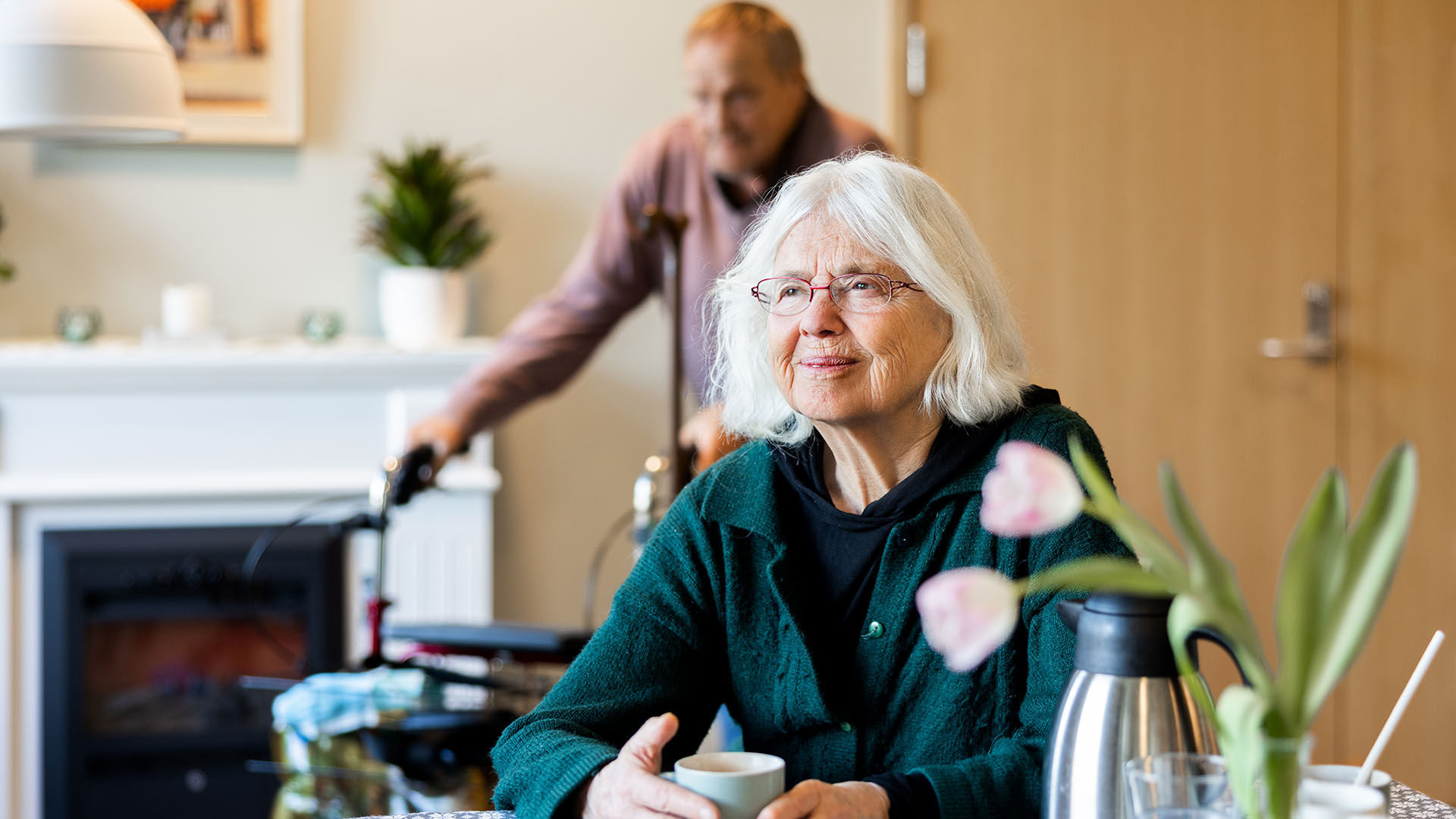 äldre kvinna sitter vid ett bord och tittar snett framåt, en äldre man i bakgrunden, en kaffekopp och tulpaner på bordet
